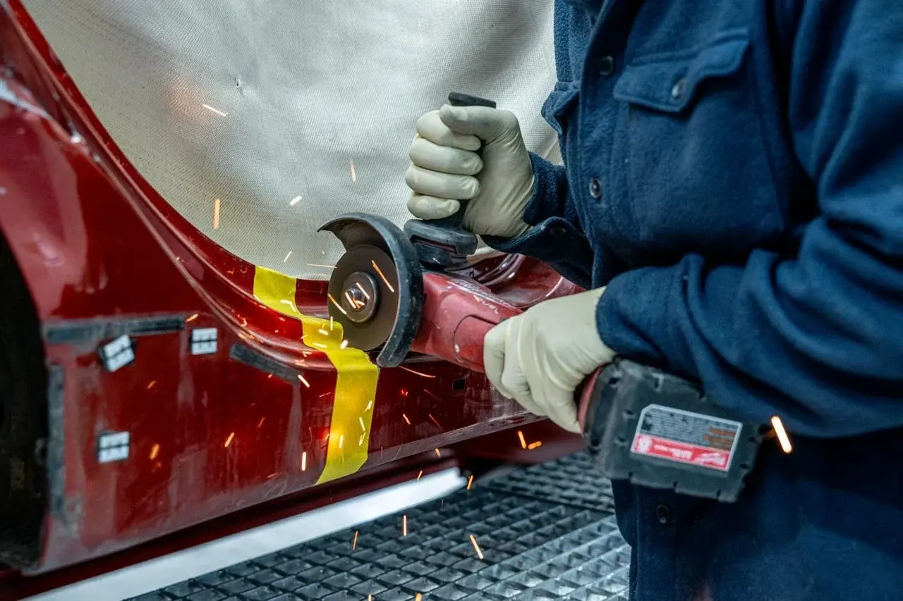 Technician in gloves grinding the lower side panel of a red car, with sparks flying past yellow tape