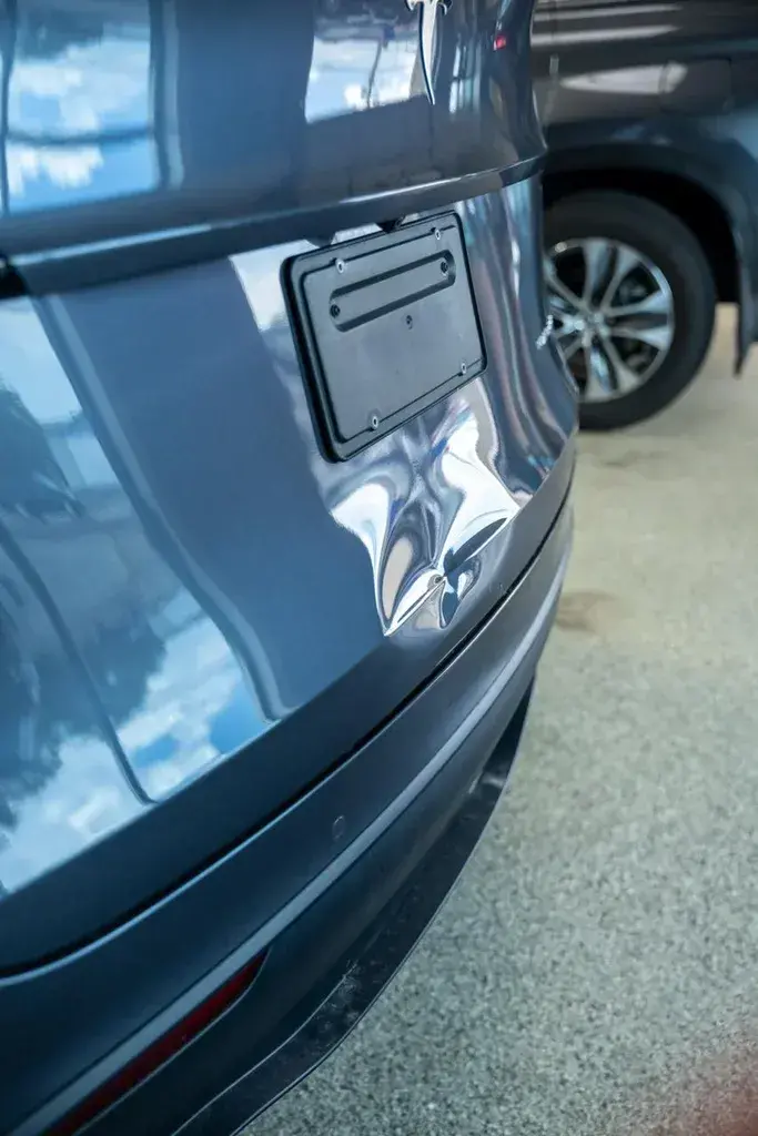 Close-up of the rear liftgate of a gray SUV with a crease dent below an empty license plate bracket, parked on a concrete floor beside another vehicle