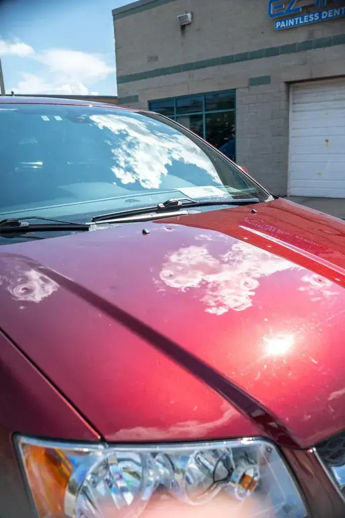 Hood of a maroon minivan with several small dents visible in the reflections of the clouds, parked outside a building with an EZ Tech Paintless Dent sign