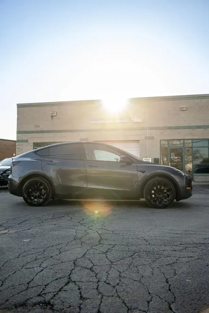 Side view of a gray crossover SUV parked on cracked asphalt in front of a building with a Paintless Dent Removal sign, with the sun shining over the roofline