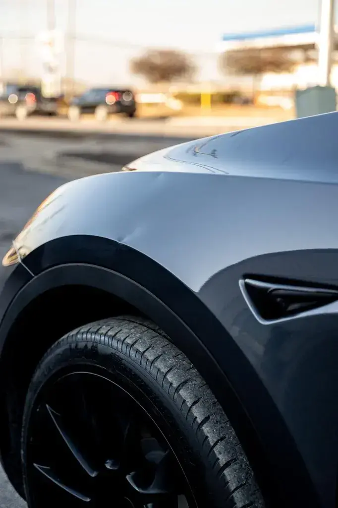 Close-up of the front fender and black wheel of a dark gray car in low sunlight, with the corner of a headlight at left and out-of-focus cars in a parking lot behind