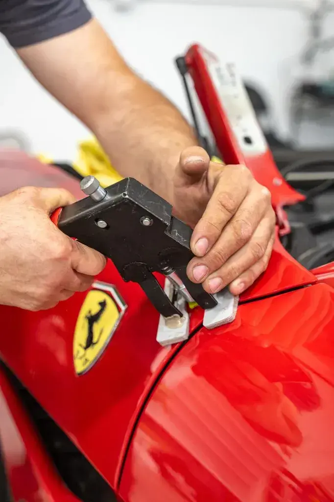 Two hands working a black glue-pull dent tool with white glue tabs on the fender of a red Ferrari, next to the yellow prancing horse badge