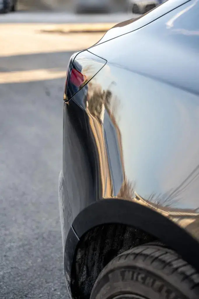 Rear quarter panel and taillight of a gray car reflecting the sky, with the rear tire and wheel arch at the bottom, parked on a sunny asphalt lot