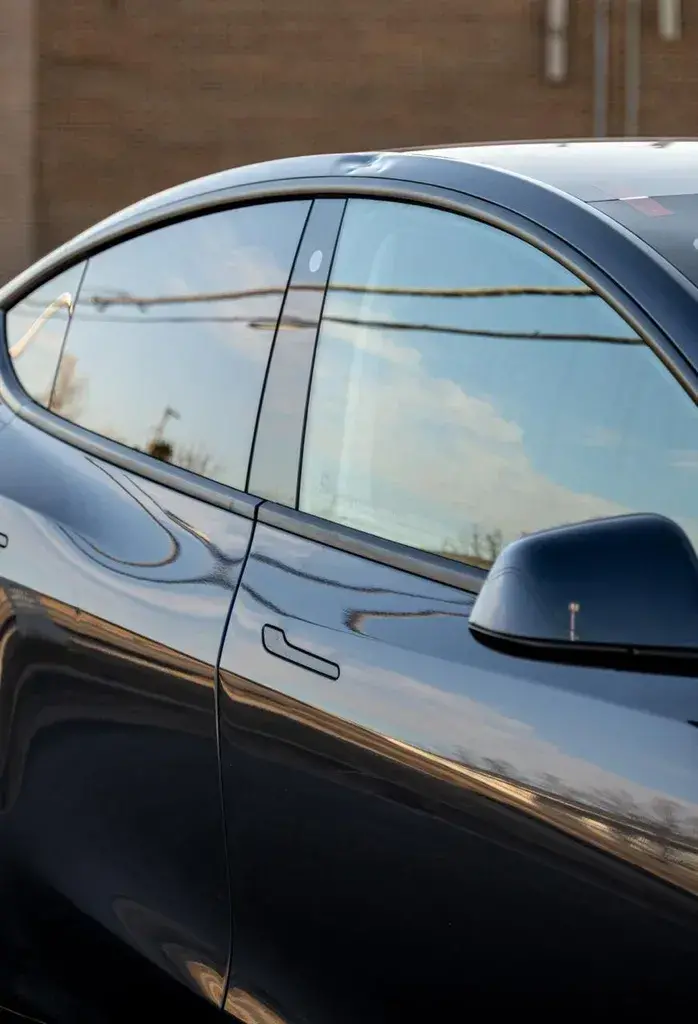 Side of a dark gray car with the front and rear doors, door handle and side mirror, reflecting a blue sky, with a brick wall behind